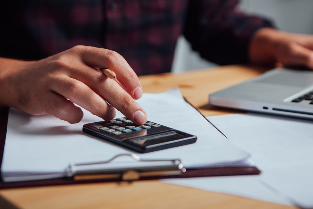 Uma mão a utilizar uma calculadora sobre documentos, com um portátil desfocado ao fundo.