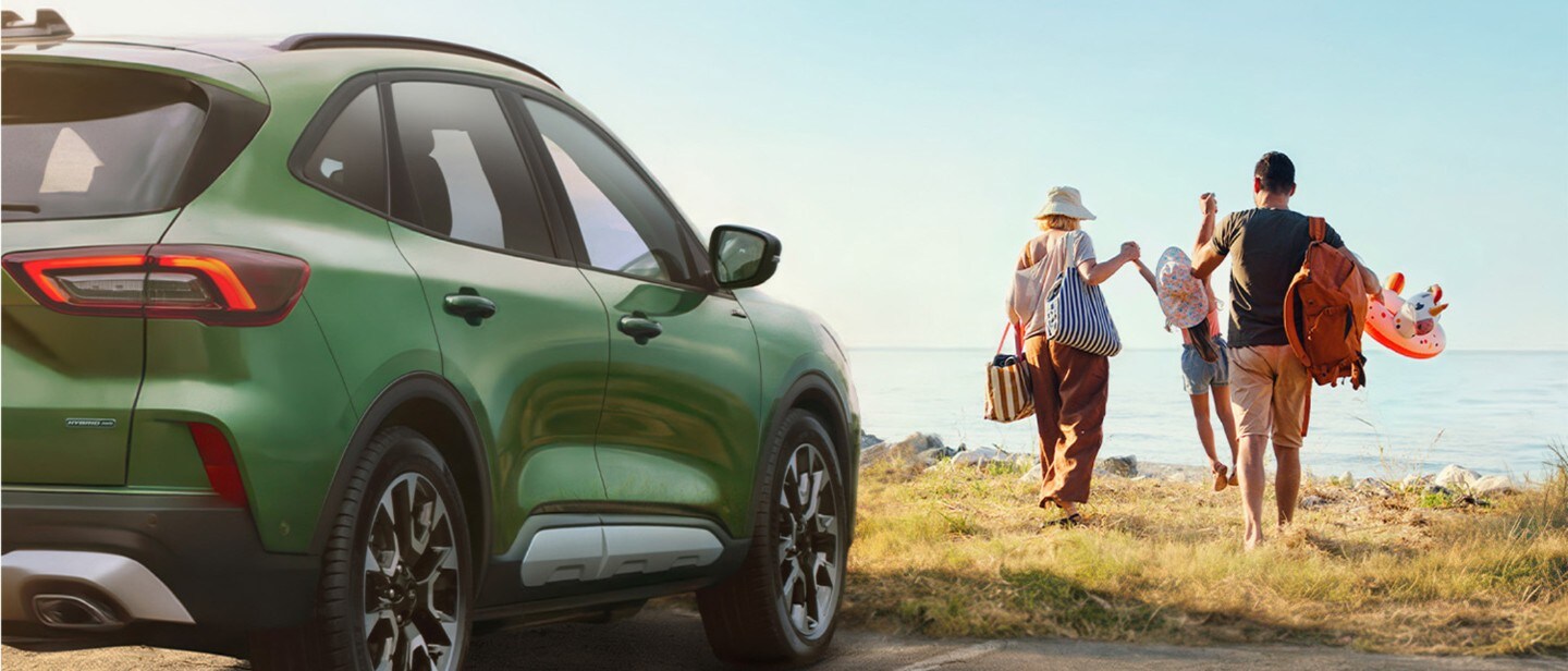 Ford Kuga verde estacionado na costa enquanto uma família caminha de mãos dadas em direção a uma praia soalheira.