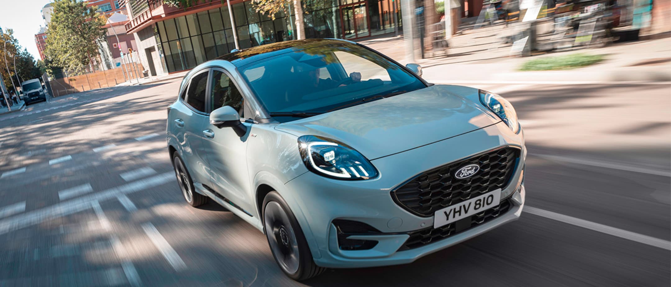 Ford Puma cinzento claro ou azul celeste com teto preto, circulando em movimento numa rua urbana ensolarada com o fundo desfocado.