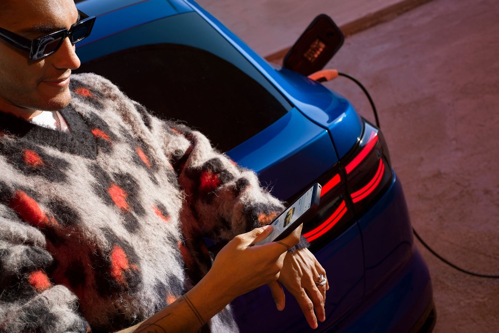 Um homem olha para o telefone enquanto se apoia no porta-malas de um Ford Capri elétrico azul.