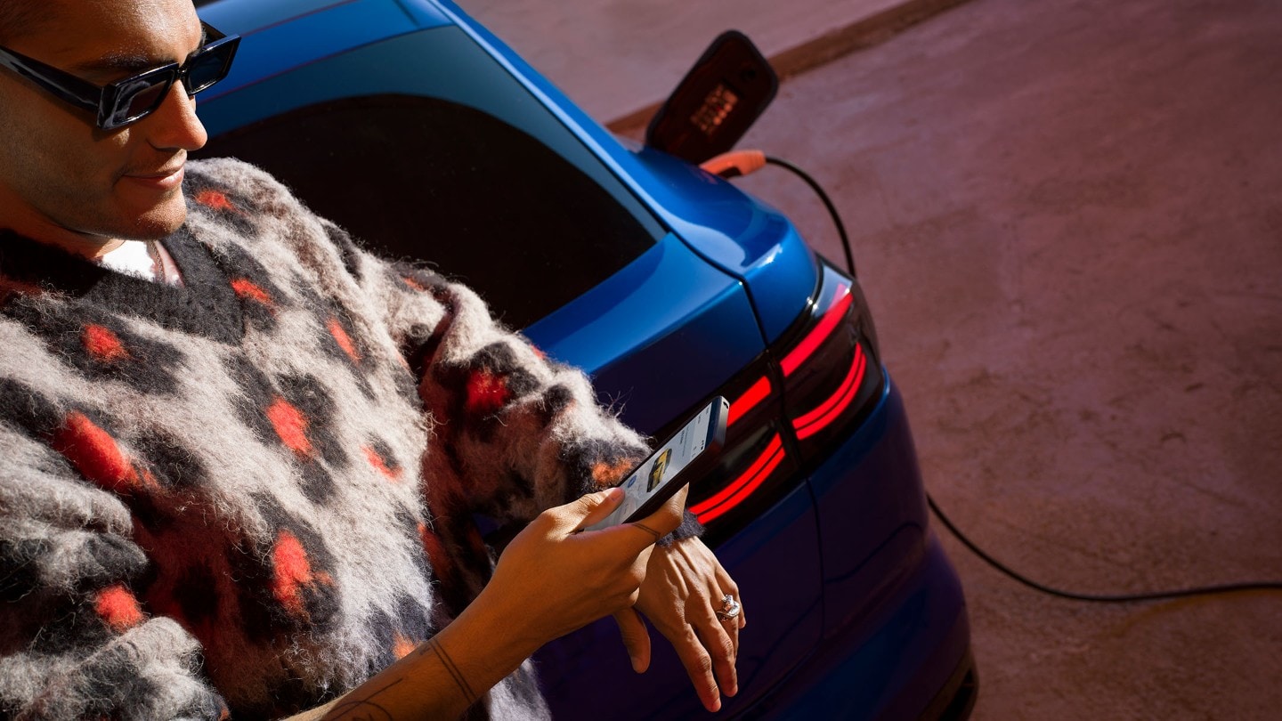Um homem olha para o telefone enquanto se apoia no porta-malas de um Ford Capri elétrico azul.