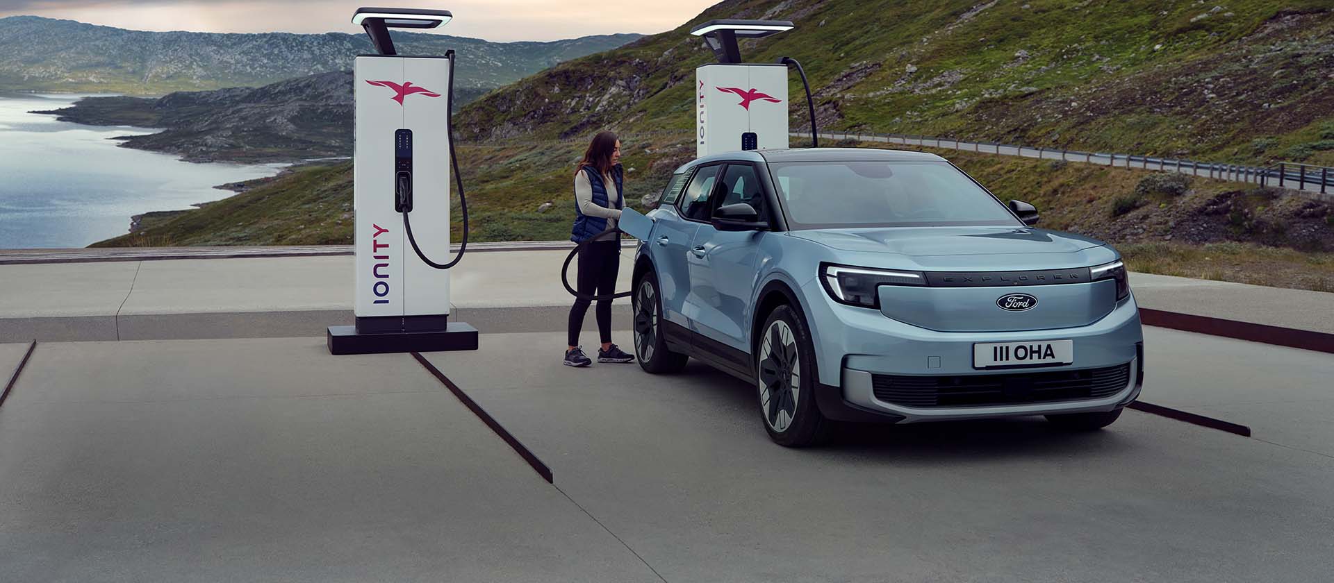 Uma mulher conecta um Ford Explorer azul, totalmente elétrico, a uma estação de carregamento pública usando um cabo. O carro está estacionado perto de montanhas e água.