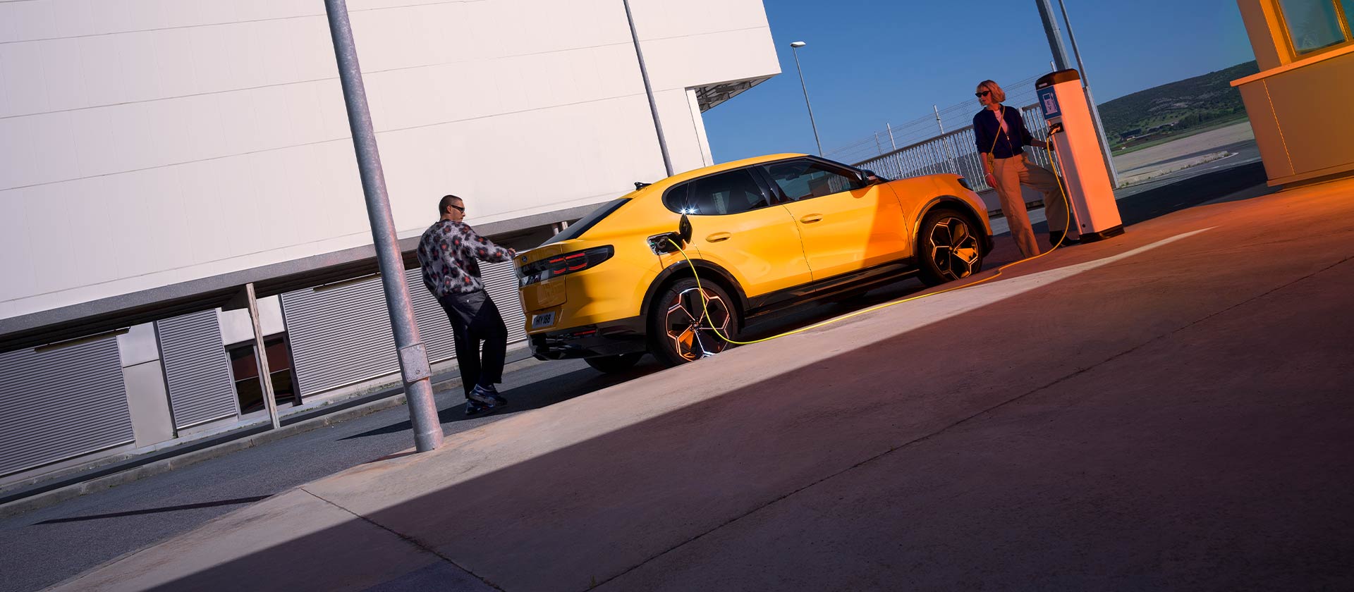 Pessoas carregando um Ford Capri EV amarelo em uma estação de carregamento pública.
