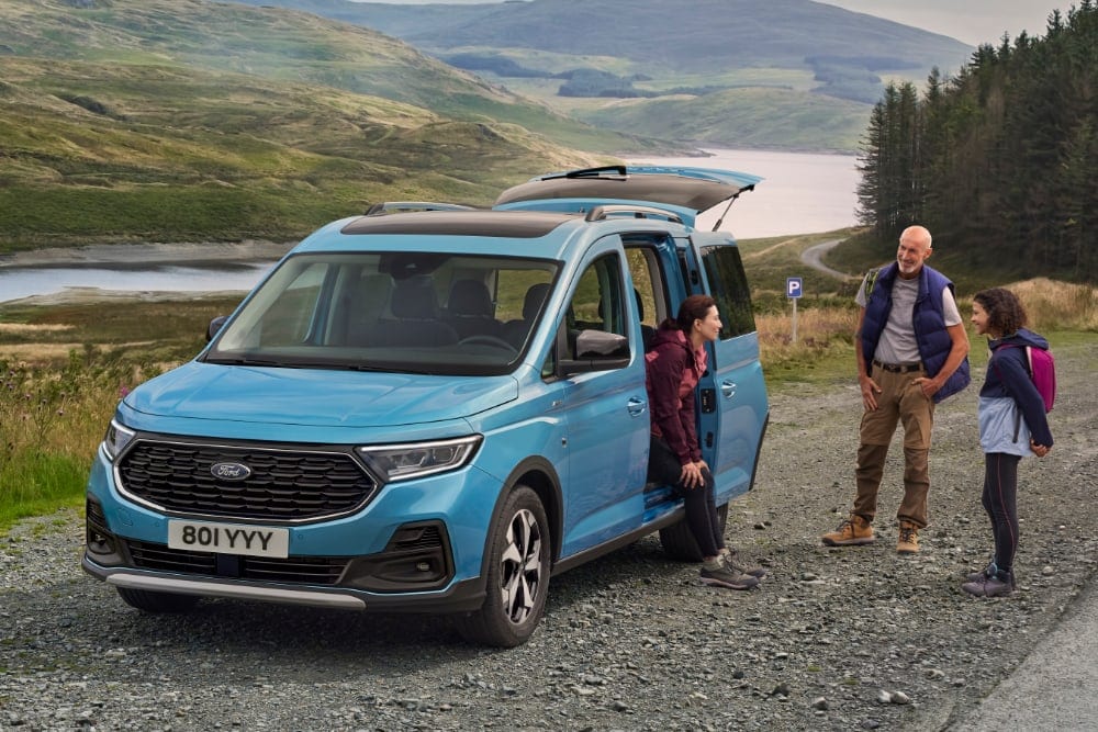 A blue Ford Tourneo Connect parked by a lake, with people getting out of the vehicle.