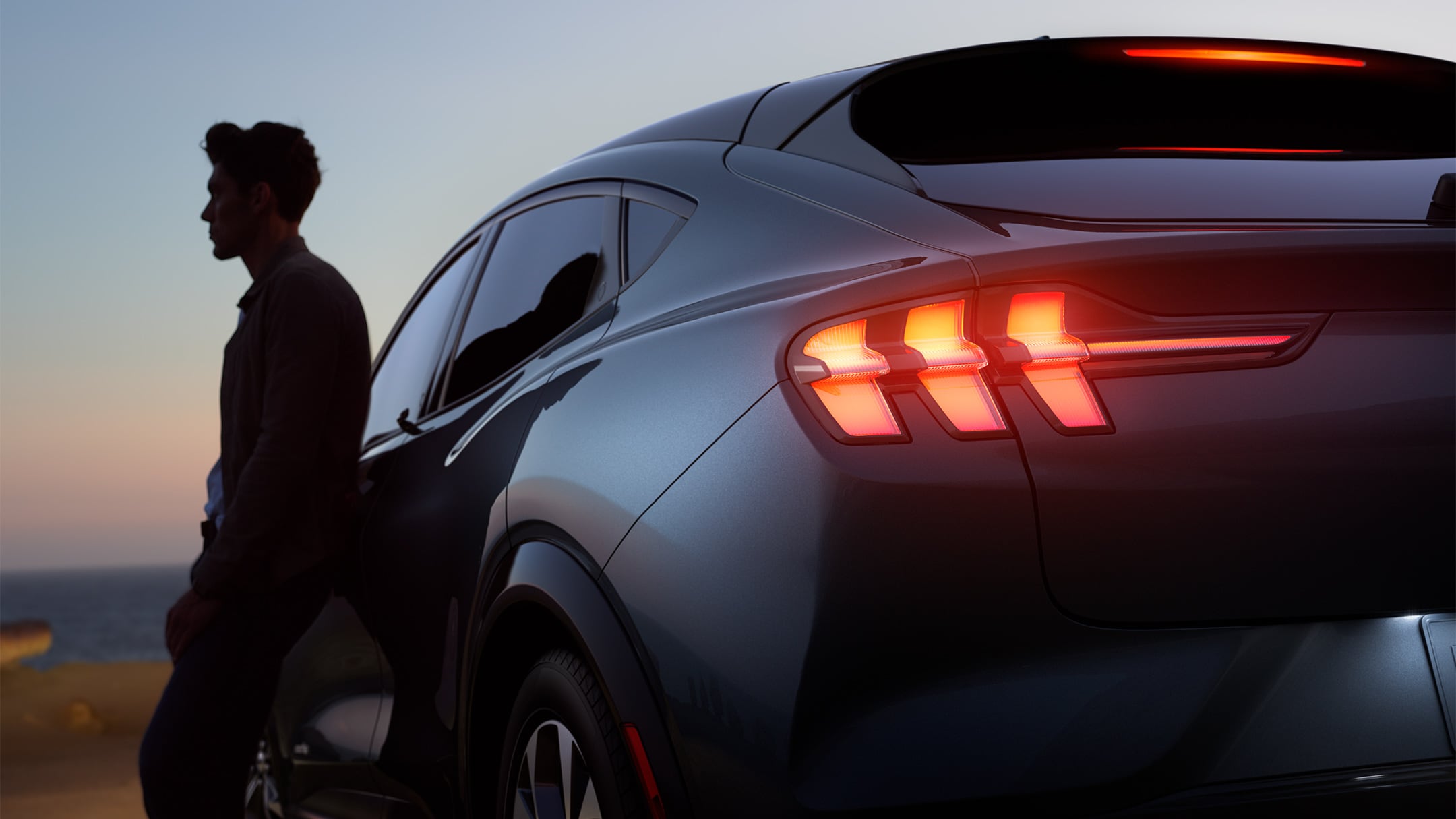 A man leaning against an All-New Ford Mustang Mach-E at sunset