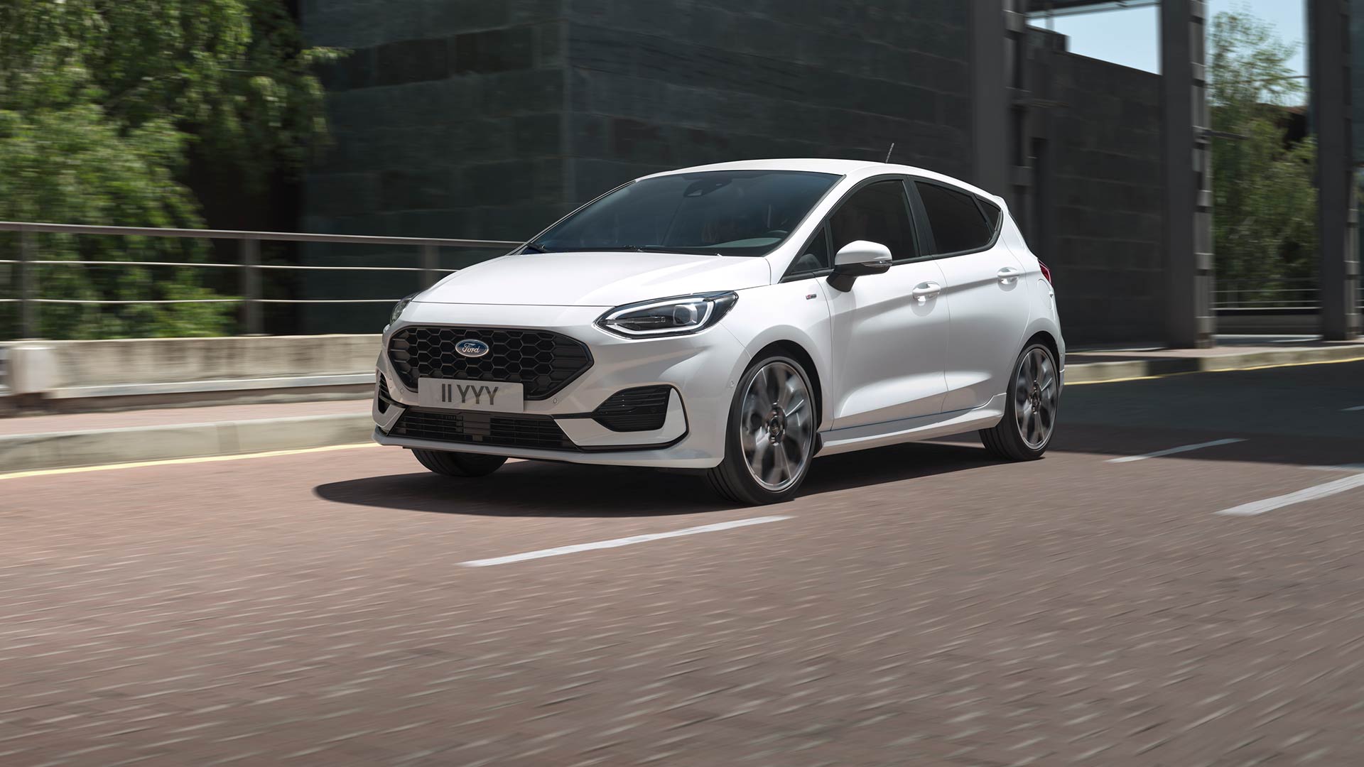 Um Ford Fiesta branco a circular numa cidade, com vegetação e edifícios ao fundo.