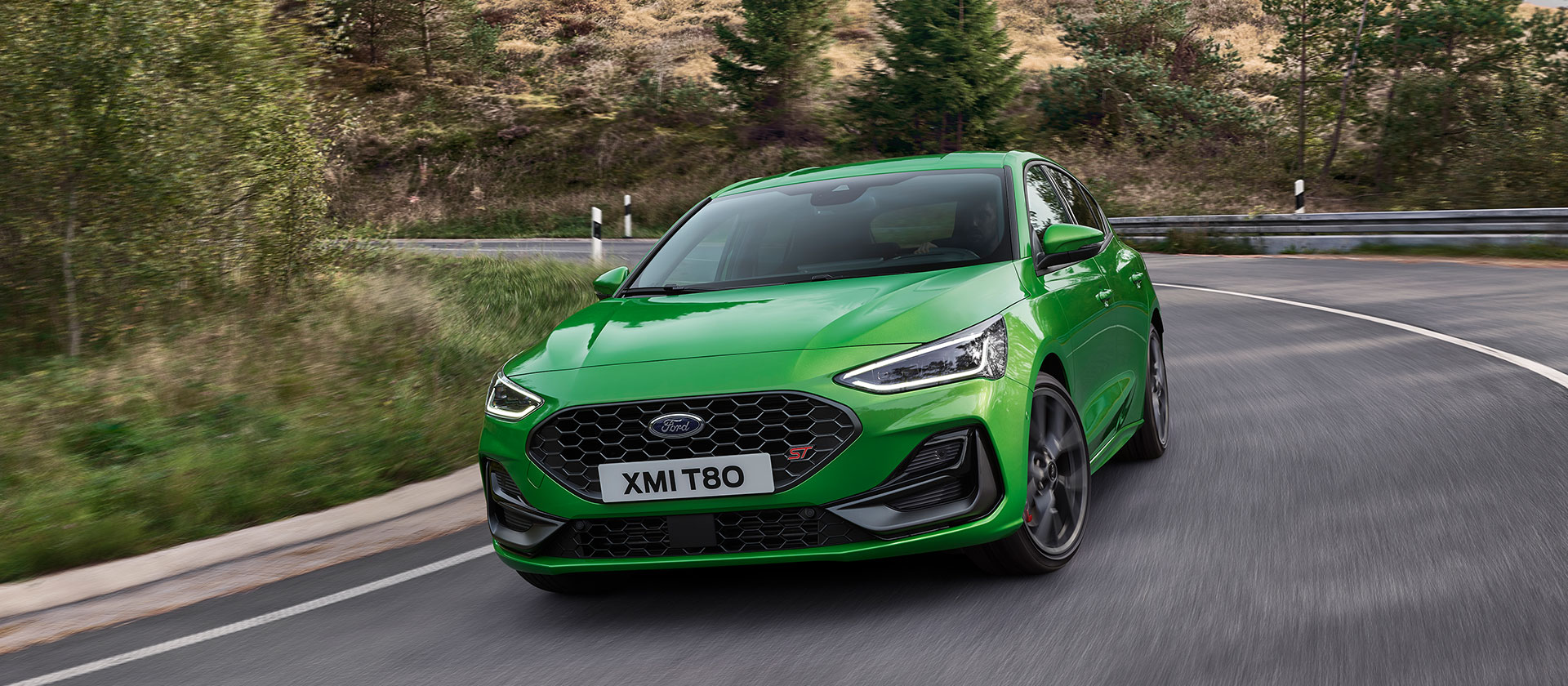 Um Ford Focus ST verde médio dirigindo em torno de uma esquina em uma estrada cênica.
