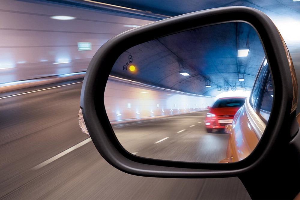 Um close-up do espelho retrovisor lateral em um túnel, mostrando um carro vermelho se aproximando no ponto cego do motorista.
