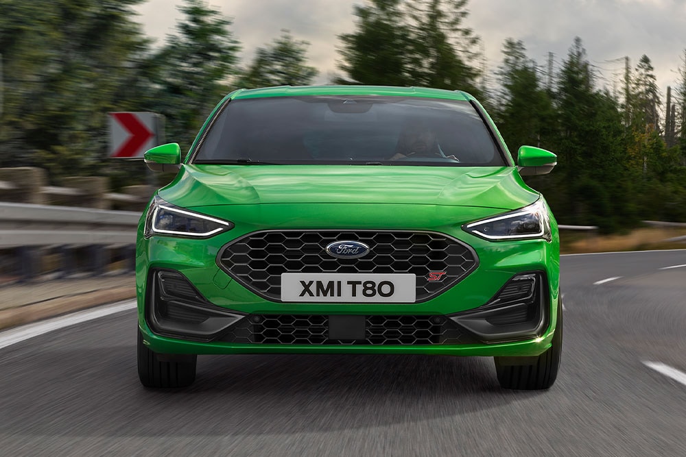 A frente de um Ford Focus ST verde médio contornando uma esquina, com árvores borradas ao fundo.