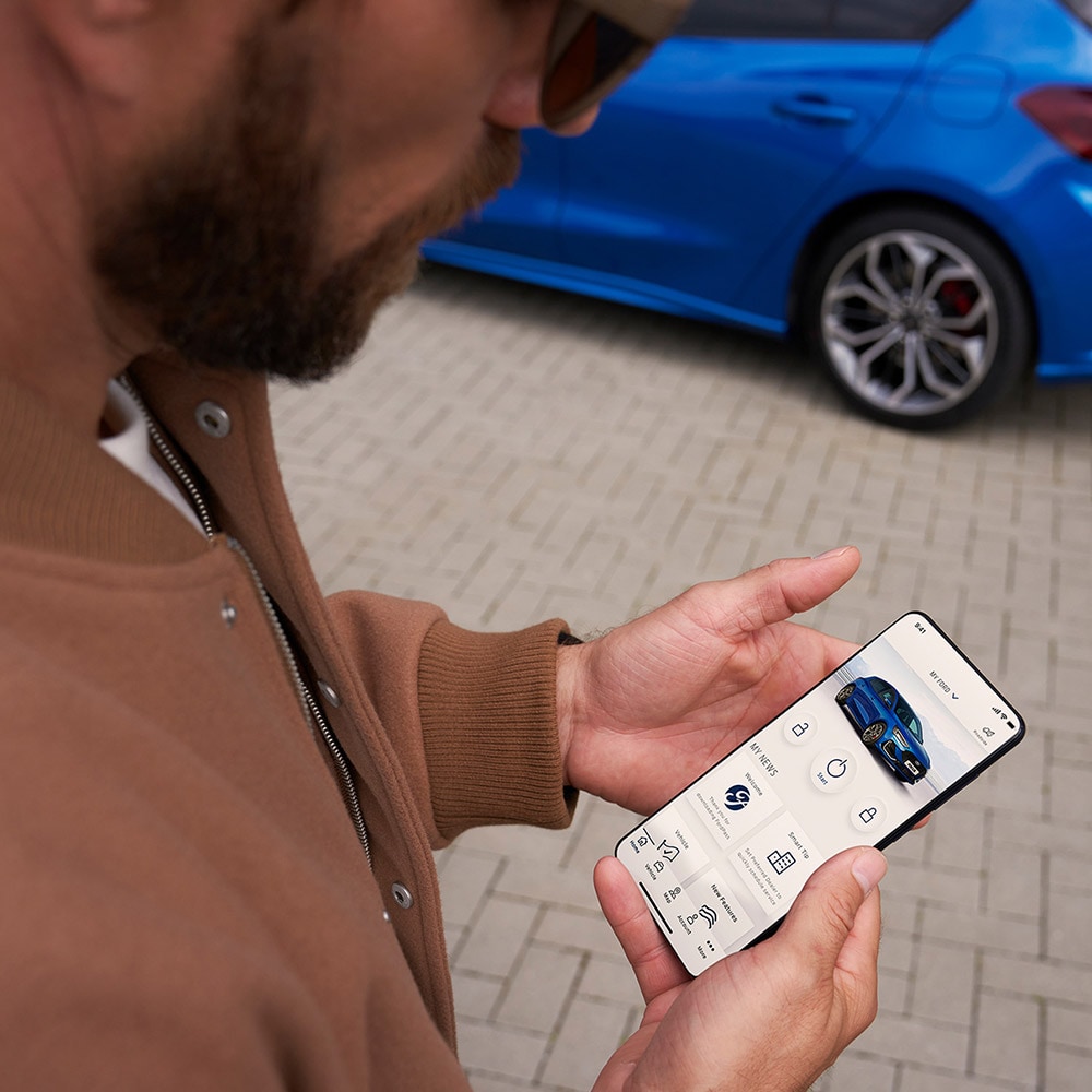 Uma pessoa segurando um smartphone exibindo o aplicativo FordPass™, com um Ford Focus azul ao fundo.