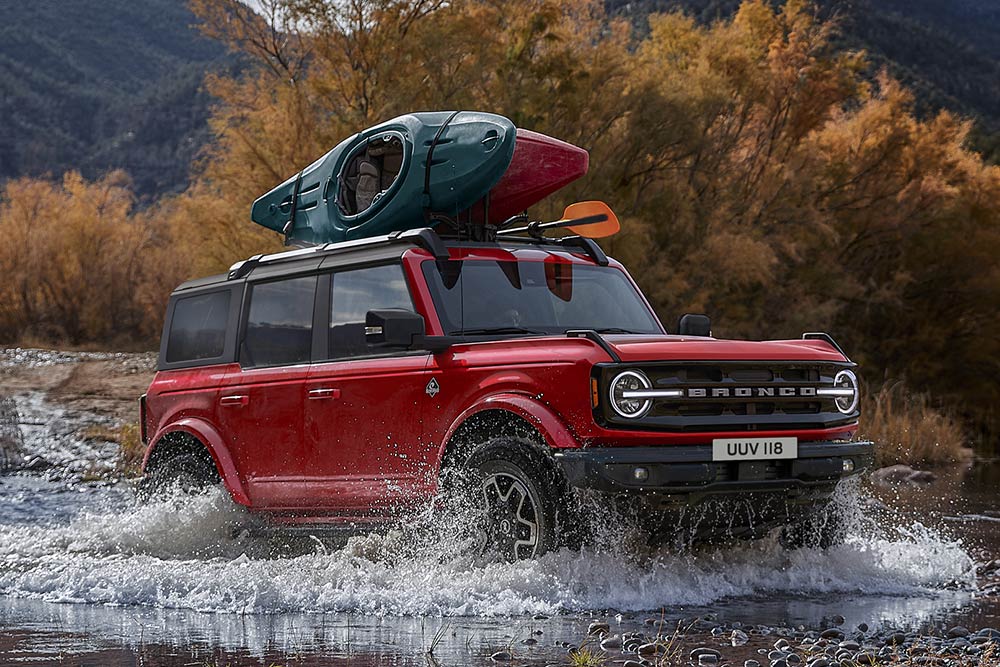 Vista da parte lateral de um Ford Bronco vermelho a atravessar um ribeiro num cenário de montanha.