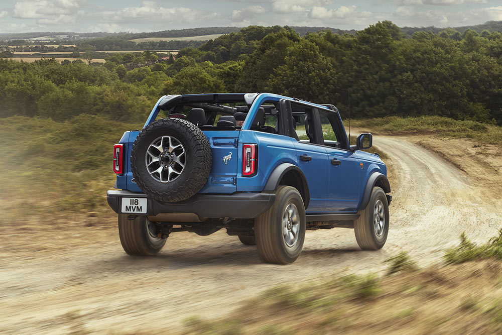 Vista da parte traseira de um Ford Bronco azul a percorrer uma paisagem campestre.