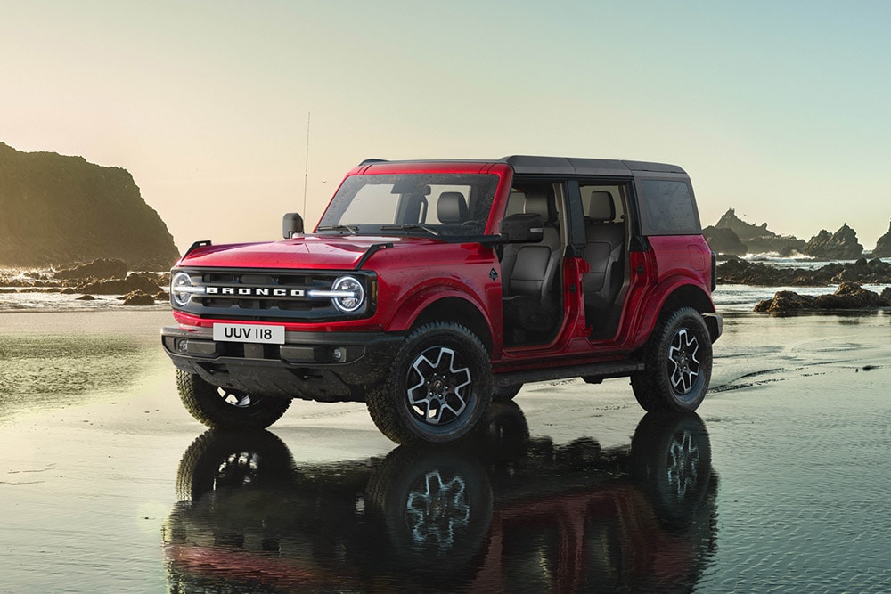 Vista da parte lateral de um Ford Bronco vermelho, com as portas removidas, estacionado numa praia de areia molhada.