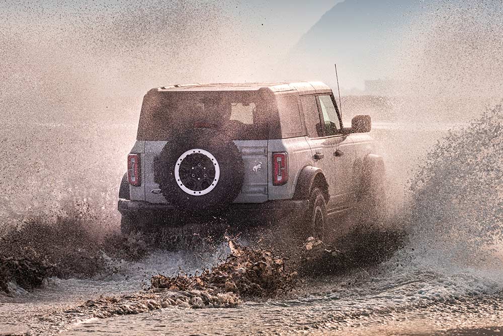 Vista da parte traseira de um Ford Bronco prateado a percorrer uma praia, provocando salpicos de água.