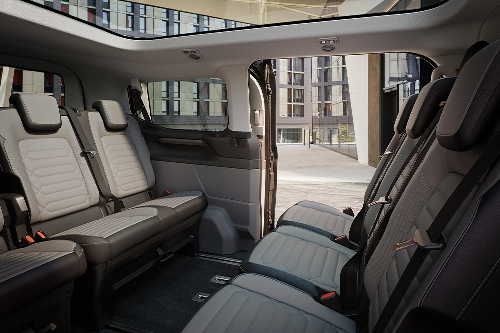 Vista do interior do Ford Tourneo Custom, a mostrar várias filas de bancos de passageiros cinzentos e o teto de abrir panorâmico.