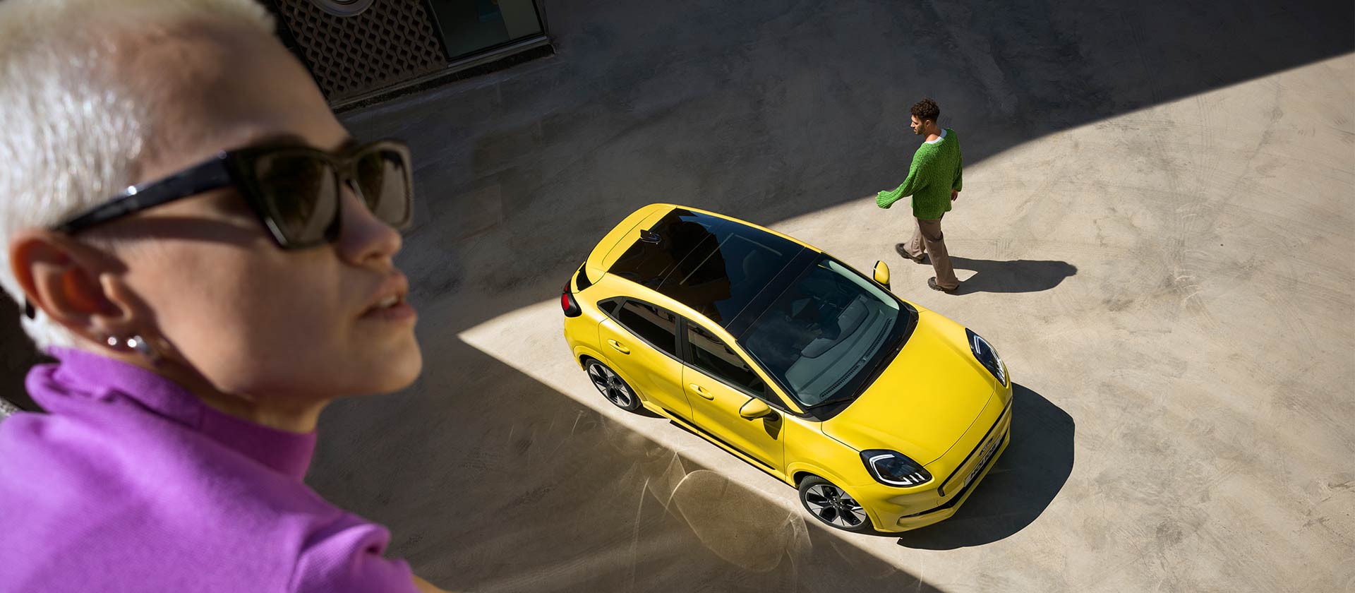 Vista aérea de um Ford Puma Gen-E® elétrico amarelo. Pessoa vestida de roxo em primeiro plano.