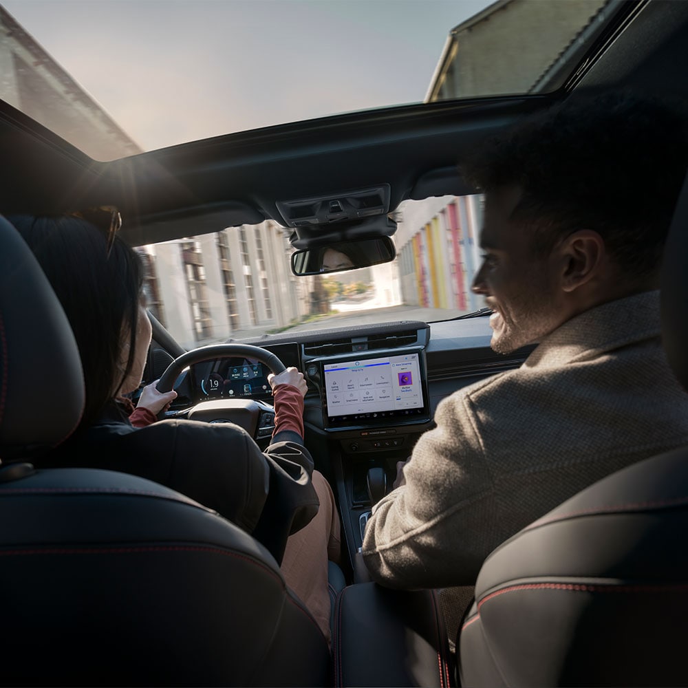 Condutor e passageiro a conversar enquanto conduzem por uma zona residencial ensolarada.