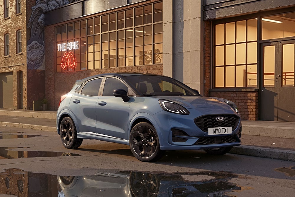 Um Ford Puma azul estacionado numa rua urbana ao pôr do sol, com poças de água a refletirem os edifícios.