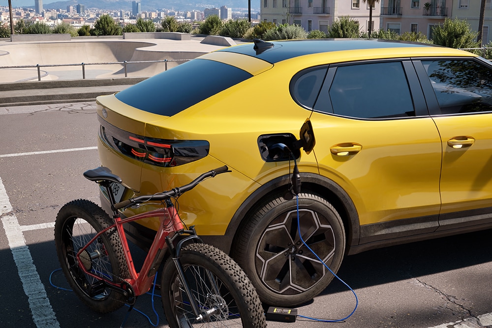 Um Ford Capri® amarelo estacionado, a carregar uma bicicleta elétrica vermelha numa paisagem urbana.