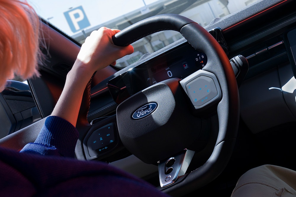 Grande plano de uma mulher a segurar o volante de um Ford Capri estacionado, mostrando também o painel de instrumentos digital.