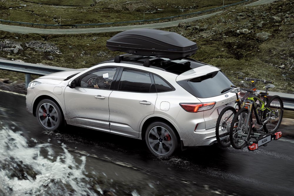 Um Ford Kuga prateado percorre uma estrada de montanha com caixa de tejadilho e suporte de bicicletas atrás.