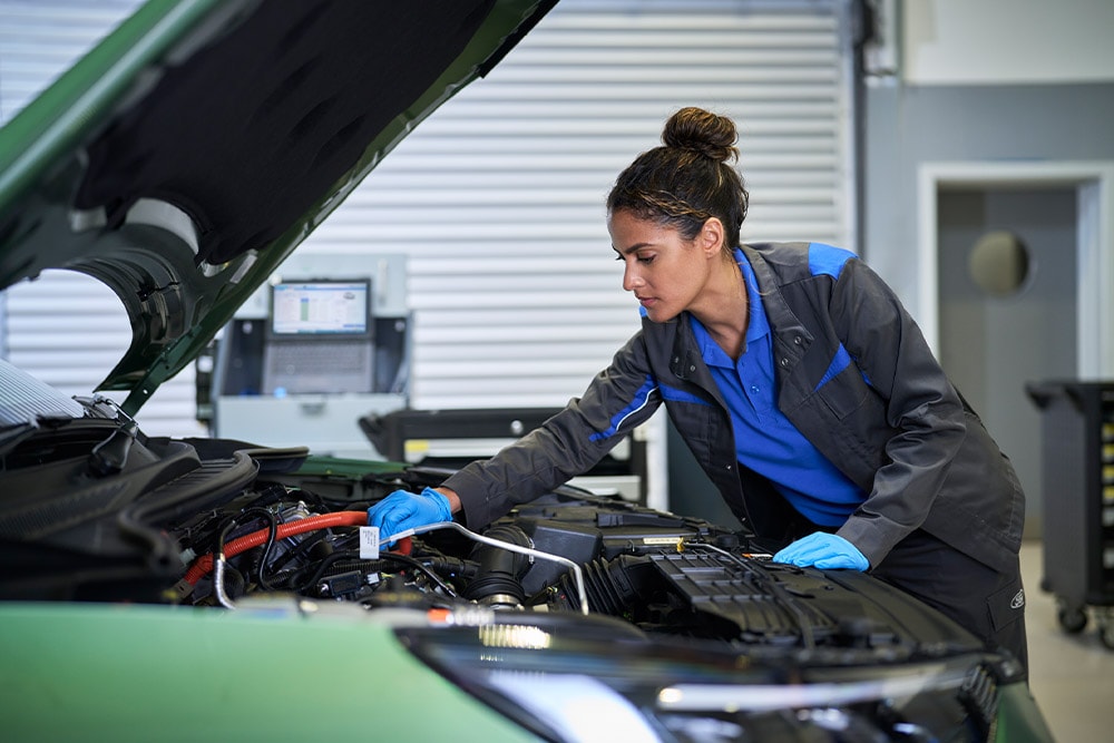 Um técnico com luvas azuis trabalha no motor de um carro verde com o capô aberto.