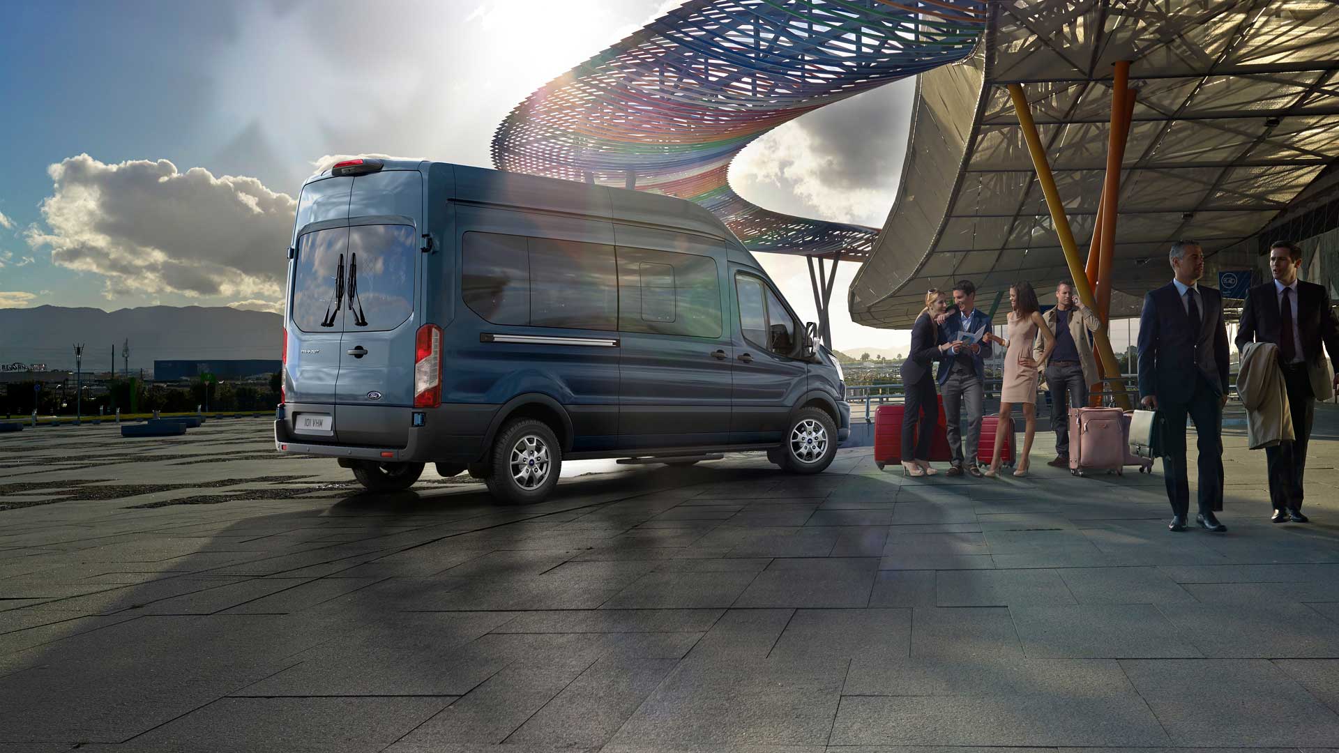 Vista de 3/4 traseira de uma Ford Transit Minibus azul estacionada num aeroporto, com pessoas e bagagens nas proximidades.