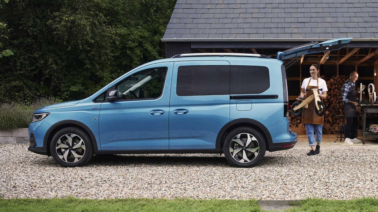 Ford Tourneo Connect in Blau. Seitenansicht, parkt vor einem Haus mit geöffnetem Kofferraum.