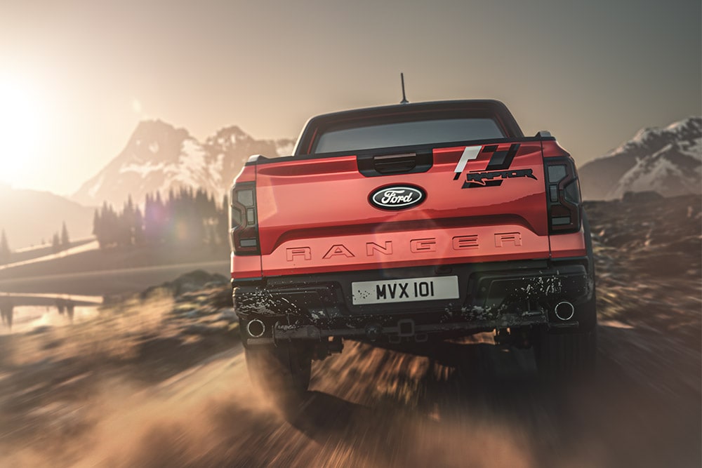 Vista traseira de um Ford Ranger Raptor vermelha numa estrada de terra batida, com montanhas ao fundo.