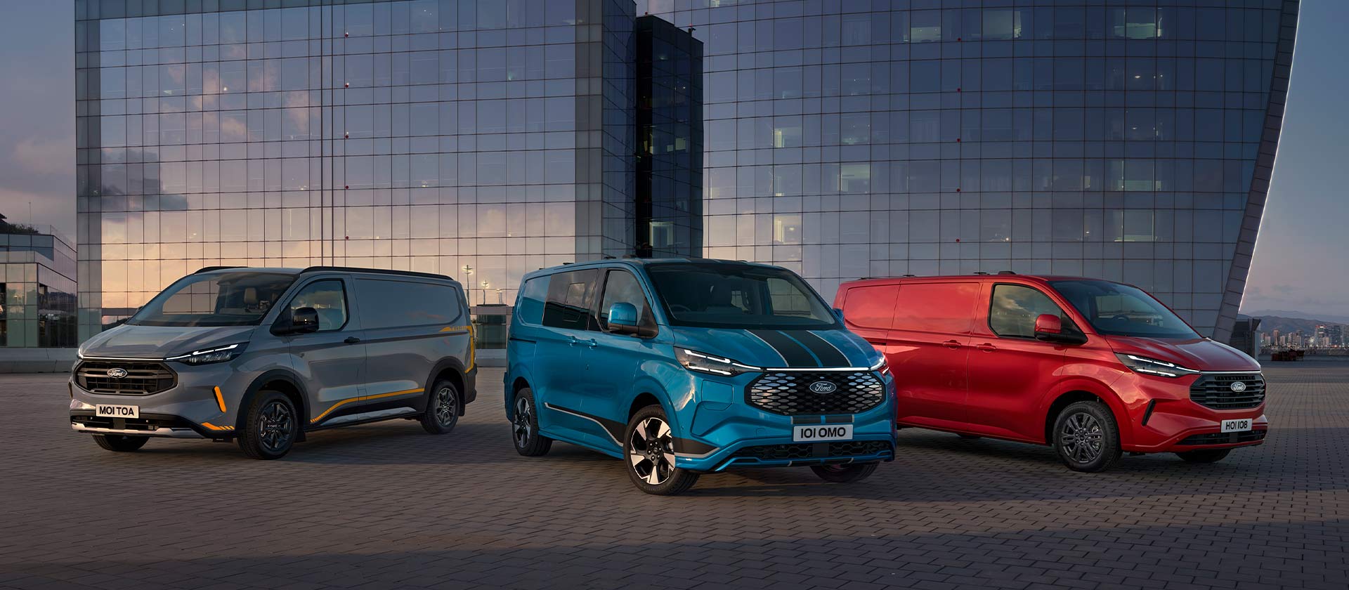 Três furgões Ford Transit Custom Sport em cinzento, azul e vermelho estacionados. 