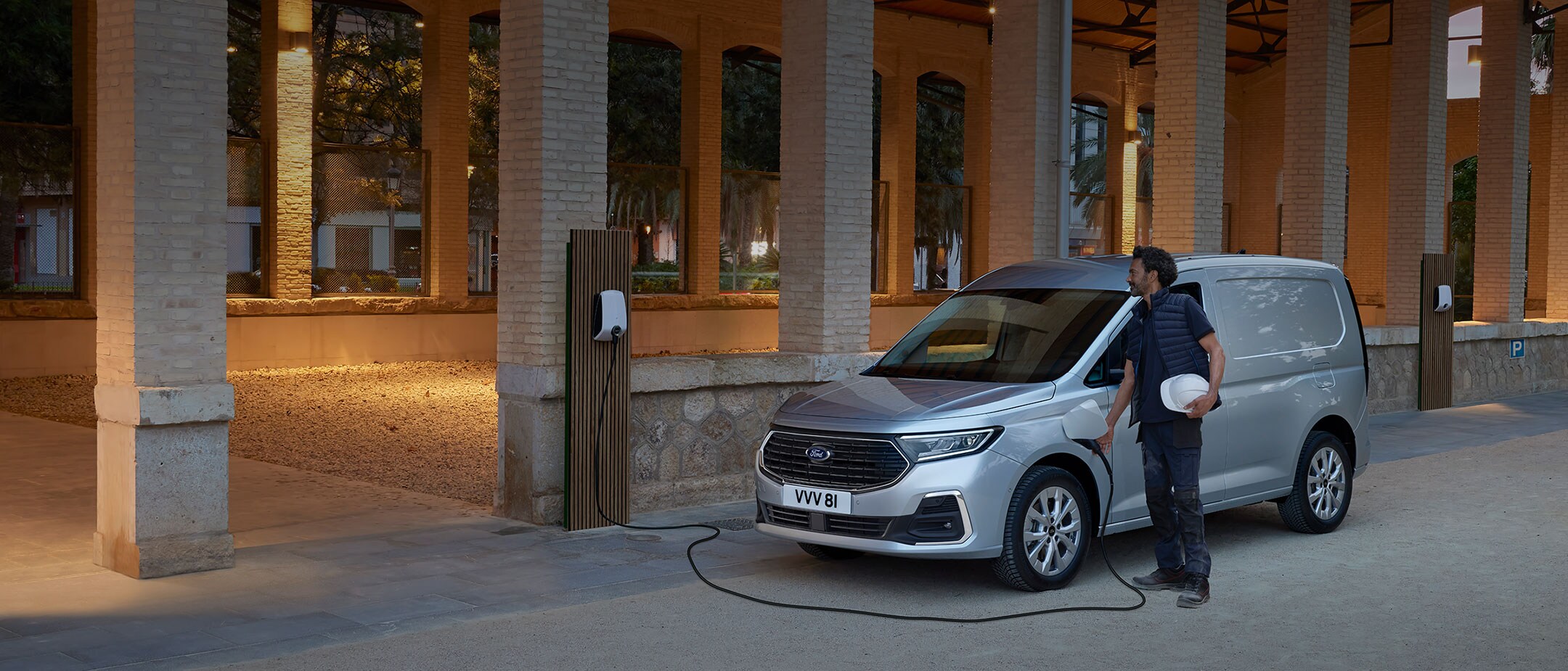 Um homem a segurar um capacete liga um cabo de carregamento de wallbox na nova Ford Transit Connect em Stardust Silver.