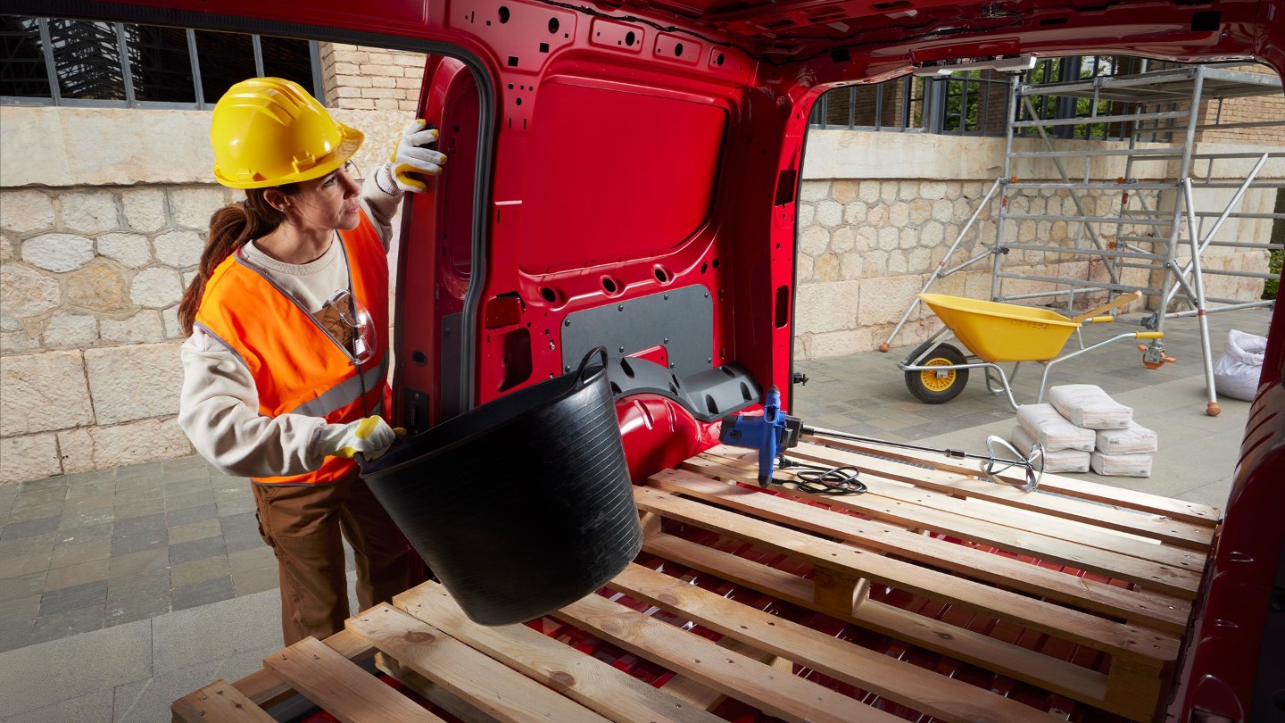 Uma mulher coloca materiais no espaço de carga da nova Ford Transit Connect em Maple Red com as portas laterais e traseiras abertas, num estaleiro de construção.