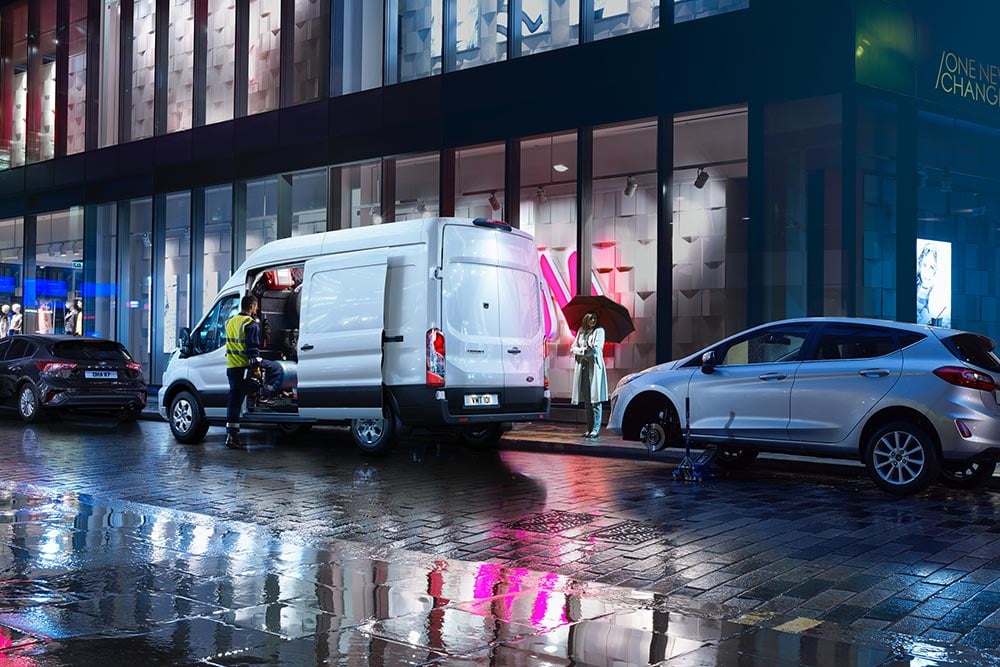 Ford Transit branca com porta lateral aberta, estacionada numa rua da cidade chuvosa, entre dois carros. 