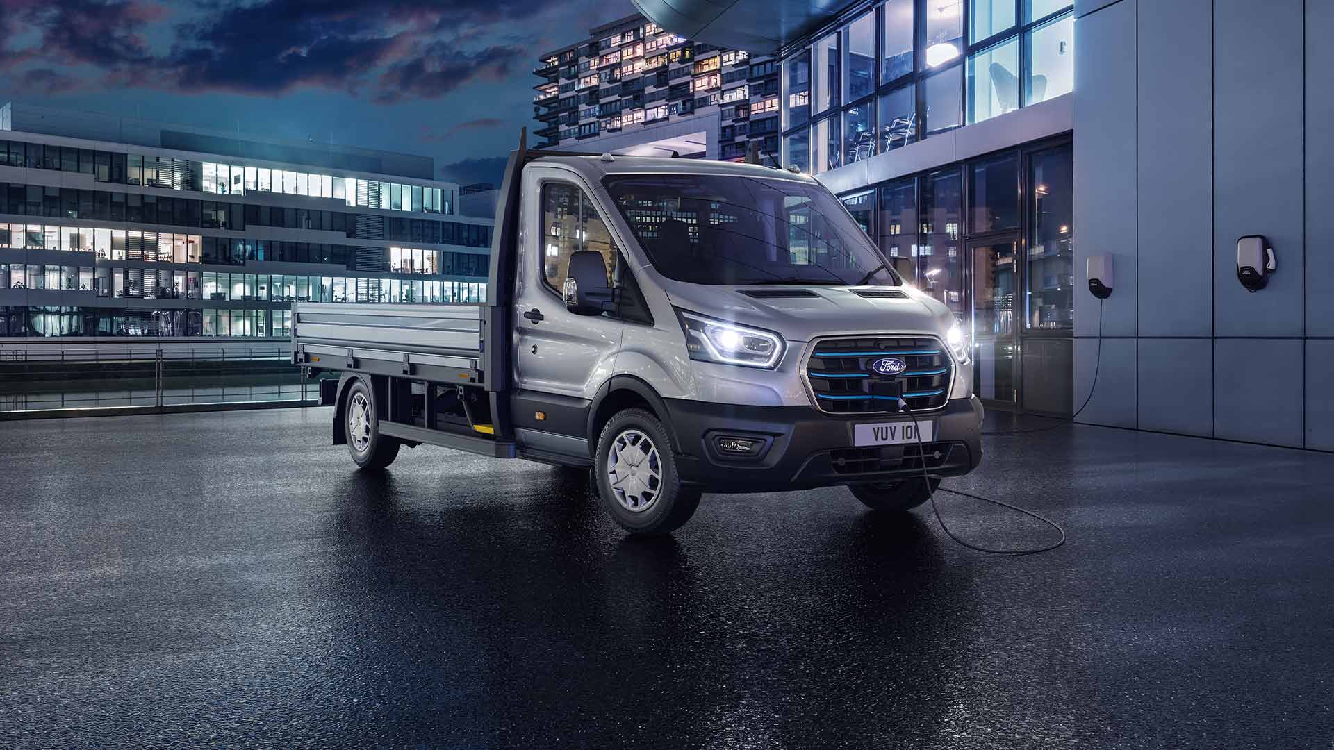 Ford Transit Chassis Cabina prateada estacionada em frente a um edifício moderno, de noite.
