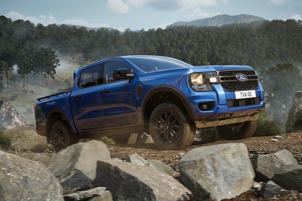 Uma pick-up Ford Ranger azul percorre uma estrada de terra batida ao longo de uma floresta, deixando um rasto de poeira.