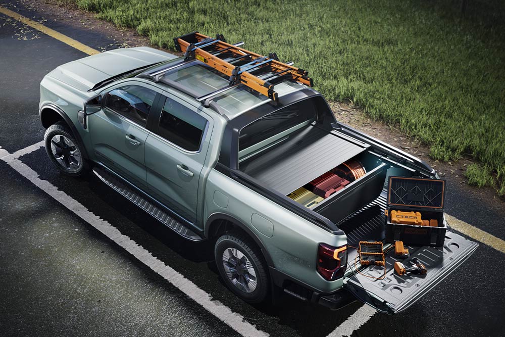 Vista traseira em ângulo alto de uma caminhonete Ford Ranger cinza, com a caçamba aberta contendo ferramentas e o bagageiro de teto, sobre uma superfície pavimentada.