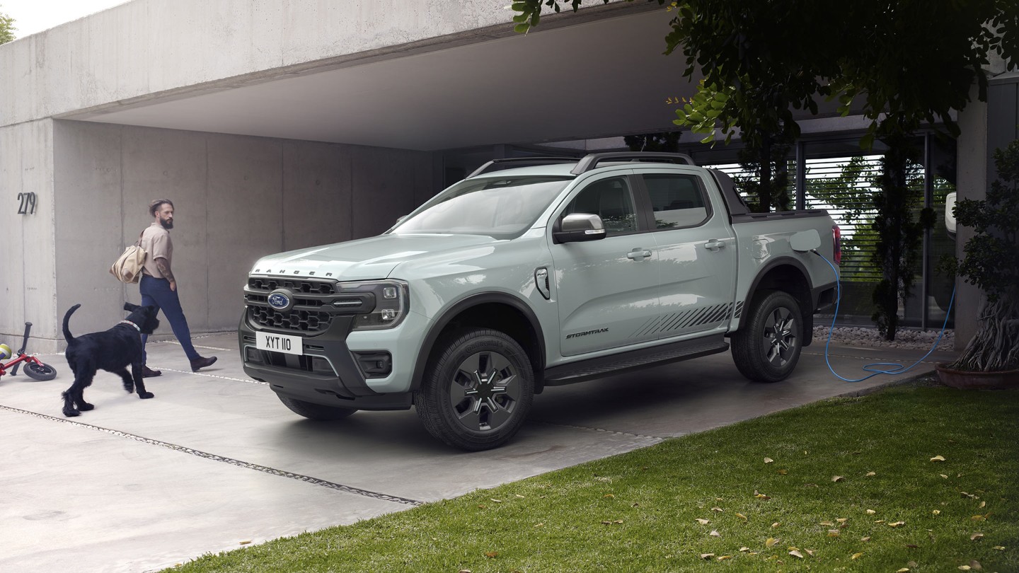 Um homem e o seu cão entram numa casa moderna, passando por um Ford Ranger Stormtrak plug-in híbrido estacionado a carregar.