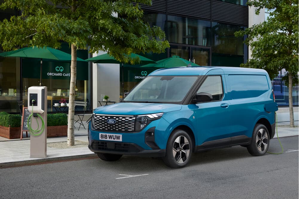 Uma Ford E-Transit Courier azul está estacionada numa rua da cidade junto a uma estação de carregamento de veículos elétrico