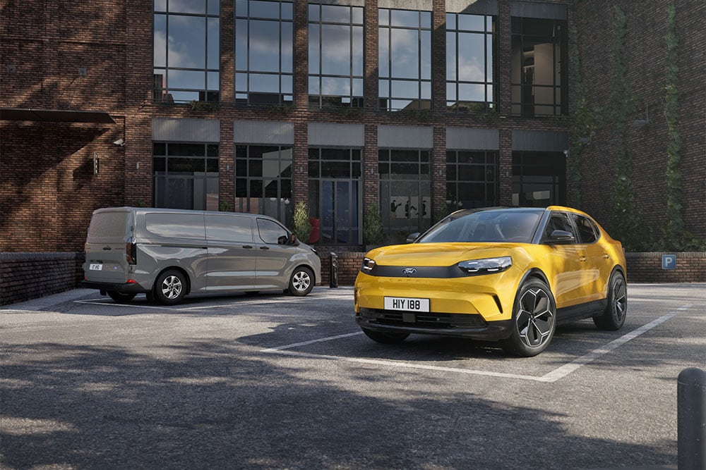 Uma carrinha Ford cinzenta e um Capri amarelo estacionados em frente a um moderno edifício de tijolo.
