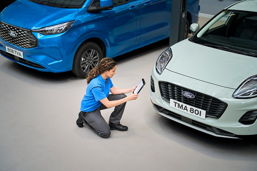 Um técnico ajoelhado entre uma carrinha Ford azul e um Ford Puma branco, a olhar para um tablet.