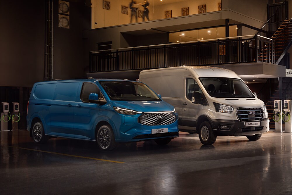 Uma Ford E-Transit Custom azul e uma Ford E-Transit prateada a carregar num parque de estacionamento.