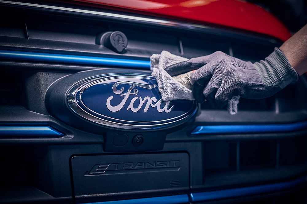 Mão com luva a tocar no emblema Ford na grelha dianteira de um veículo.