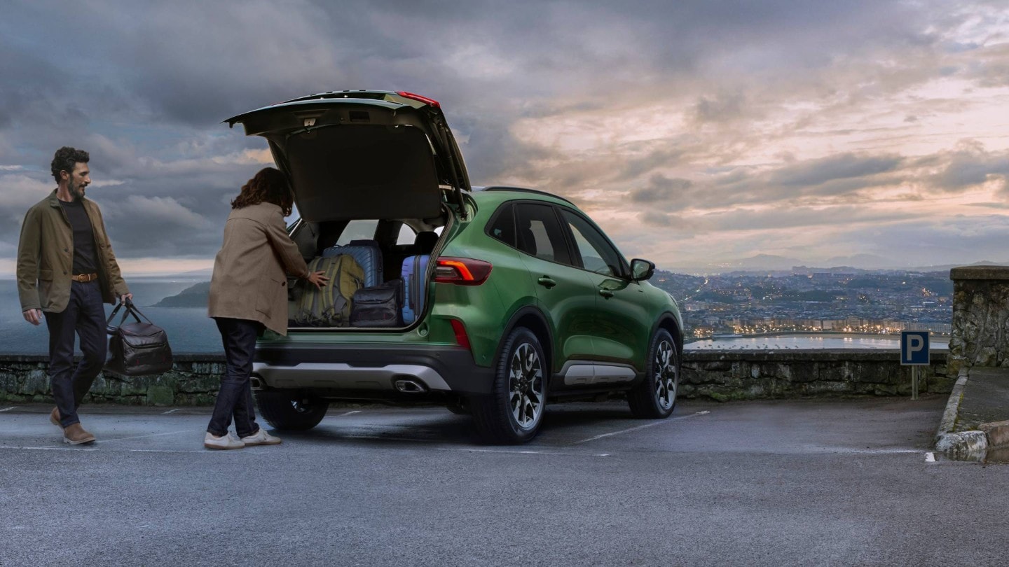 Um homem e uma mulher a colocar bagagem na bagageira de um SUV verde estacionado Um homem e uma mulher a colocar bagagem na bagageira de um SUV verde estacionado